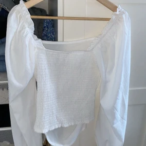 Vit offshoulder blus - Säljer en stilren vit offshoulder blus med långa ärmar. Blusen har en smockad detalj vid bysten och är perfekt för en chic look.
