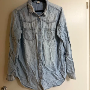 Ljusblå jeansskjorta - Säljer en ljusblå jeansskjorta med knappar och två bröstfickor. Skjortan har långa ärmar och en klassisk krage. Perfekt för en avslappnad stil.