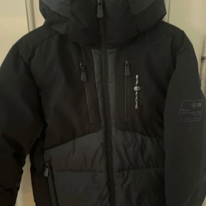 Svart jacka från Sail Racing - Säljer en svart jacka från Sail Racing med dragkedja och logotyp på bröstet och ärmen. Jackan har en hög krage och är perfekt för kalla dagar. Den är designad för att klara tuffa väderförhållanden.