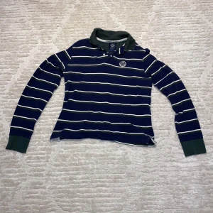 Randig pikétröja från North Sails - Säljer en snygg randig pikétröja från North Sails i blått och grönt med vita ränder. Tröjan har en klassisk krage och knappar framtill. Perfekt för en avslappnad stil.