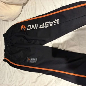 GASP - Säljer ett par svarta träningsbyxor från GASP med orange detaljer och tryck. Byxorna har elastisk midja och är perfekta för gymmet. De har en cool design med GASP-logga på sidan.