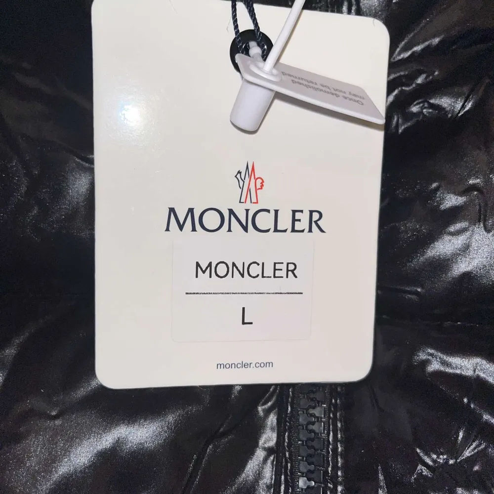 Snygg svart dunjacka från Moncler med glansig finish och dragkedja framtill. Jackan har en klassisk pufferdesign och Moncler-logga på ärmen. Perfekt för kyliga dagar.. Takit.