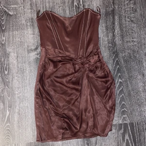 Brun klänning från Zara - Snygg brun klänning från Zara i satin med en elegant knutdetalj framtill. Klänningen har en axelbandslös design och en figurnära passform som framhäver silhuetten. Perfekt för en kväll ute eller en speciell tillställning.