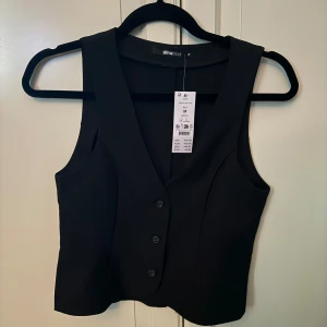 Svart väst från Gina Tricot - Snygg svart väst från Gina Tricot med knappar framtill. Perfekt för att lägga till en stilren touch till din outfit. Västen har en v-ringad design och är i en klassisk svart färg.