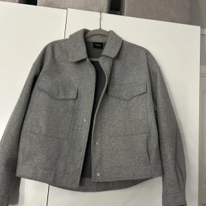 Grå jacka från Vero Moda - Snygg grå jacka från Vero Moda med en klassisk krage och knappar framtill. Jackan har två stora fickor på bröstet och en stilren design som passar perfekt för höst och vår.