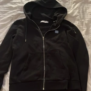 Svart hoodie från Les Deux - Snygg svart hoodie från Les Deux med dragkedja och broderad logga på bröstet. Perfekt för en avslappnad stil med praktiska fickor och huva.