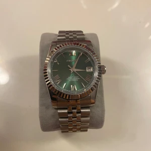 Seiko Klocka - Snygg Seiko-klocka med grön urtavla och datumvisning. Den har en silverfärgad boett och länkarmband i rostfritt stål. Perfekt för den som vill ha en stilren och tidlös accessoar. Custom gjord.