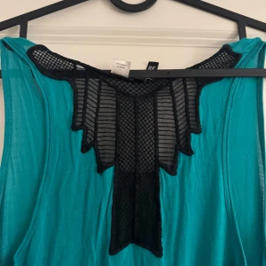 Turkos långklänning med svart spetsdetalj - Snygg turkos långklänning med unik svart spetsdetalj vid halsringningen. Perfekt för en somrig look med sin ärmlösa design och luftiga passform.