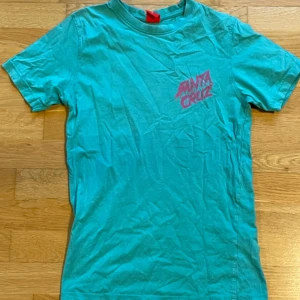 Turkos t-shirt från Santa Cruz - Cool turkos t-shirt från Santa Cruz med ett färgglatt tryck av en skateboard och monster på baksidan. Framsidan har en liten rosa logga. Perfekt för en avslappnad stil! 