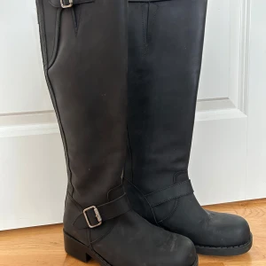 Svarta stövlar från Johnny Bull - Snygga svarta stövlar i skinn med dragkedja längs baksidan och dekorativt spänne vid ankeln. Perfekta för höst och vinter med en robust sula för bra grepp. Storlek 37. Kan mötas upp i Stenungsund eller Göteborg. 