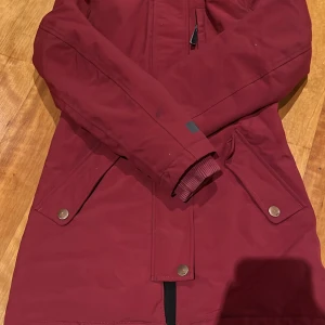 Vinröd jacka från Molo - Säljer en snygg vinröd parkajacka från Molo med fuskpäls på huvan. Jackan har dragkedja och knappar framtill samt praktiska fickor. Perfekt för kyliga dagar!
