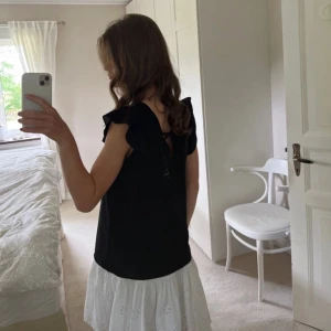 Svart ärmlös topp med volangdetaljer - Snygg svart ärmlös topp med volangdetaljer vid axlarna och en v-ringad design. Perfekt för en stilren look.