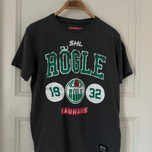 Grå Rögle t-shirt från Bauhaus - Snygg grå t-shirt med Rögle-tryck i grönt och vitt. Perfekt för hockeyfans! T-shirten har korta ärmar och är gjord i mjukt material. Bauhaus-logga syns tydligt på framsidan.