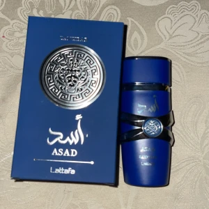 Asad Eau de Parfum från Lattafa - Elegant parfym i en stilren blå flaska med silverdetaljer. Förpackningen har ett dekorativt mönster och arabiska inskriptioner. Perfekt för den som söker en unik doftupplevelse.
