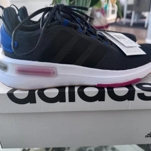 Svarta Adidas sneakers - Snygga svarta Adidas sneakers med vita sulor och blå detaljer. Skorna har en modern design med snörning och är perfekta för en sportig look. De har en bekväm passform och är idealiska för vardagsbruk.