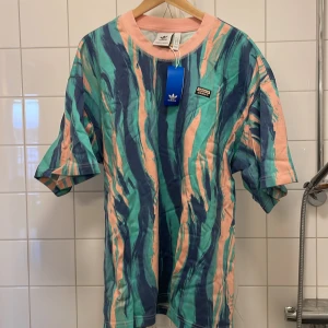 Flerfärgad t-shirt från Adidas (sällsynt) - Säljer en cool flerfärgad t-shirt från Adidas med ett abstrakt mönster i blått, grönt och rosa. T-shirten är tillverkad i tjockt skönt material och har korta ärmar och en rund halsringning. Perfekt för en avslappnad stil. Oanvänd i förpackning och ettiket kvar