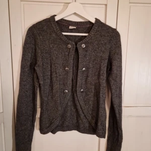 Grå stickad kofta från Esprit strl M - Fin grå stickad kofta med knappar framtill från Esprit. Stängs med dn hyskehake eller bärs öppen. Fin design och ribbade detaljer vid ärmslut och nederkant.                                          Koftan är av Ullmix och kan upplevas lite stickig för den som är känslig för stickiga plagg.  Sparsamt anvönd. 