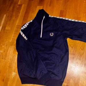 Mörkblå tröja med dragkedja från Fred Perry - Unik mörkblå halfzip från Fred Perry. Tröjan har vita detaljer längs ärmarna med mönster. Perfekt för en stilren look. Skick 10/10, Size M men passar även S