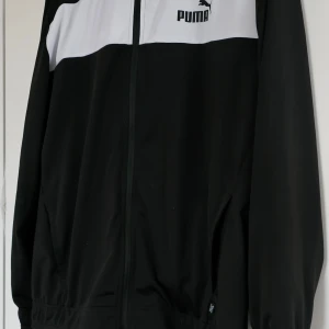 Svart och vit träningsoverall från Puma - Snygg svart och vit träningsoverall från Puma, två delar med fickorl. Perfekt för en sportig look med långärmade ärmar och klassisk design. Logotypen är tryckt på bröstet.