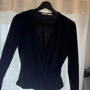 Svart omlottopp från NA-KD - Snygg svart omlottopp från NA-KD i storlek S. Toppen har långa ärmar och en stilren design som passar perfekt till olika tillfällen. Perfekt för dig som vill ha en mångsidig och elegant topp i garderoben.