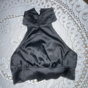 Svart axelbandslös topp - Elegant svart axelbandslös topp med knytning i ryggen. Tillverkad i ett glansigt material som ger en lyxig känsla. Perfekt för en kväll ute. Nyskick