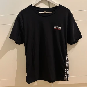 Svart t-shirt från Moschino - Snygg svart t-shirt från Moschino med logotyp på bröstet. Den har en cool detalj med text längs sidan. Perfekt för en stilren look.