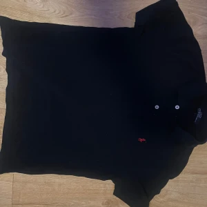 Svart kortärmad pikétröja med broderad logga - Snygg svart pikétröja från Ralph Lauren med korta ärmar och klassisk krage. Tröjan har en broderad röd logga på bröstet och knappar vid halsen. Perfekt för en stilren look.