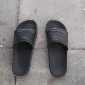 Svarta sandaler - Säljer ett par svarta sandaler i syntetmaterial. Perfekta för sommaren och enkla att glida i när du är på språng. De har en stilren design med två ränder över foten.