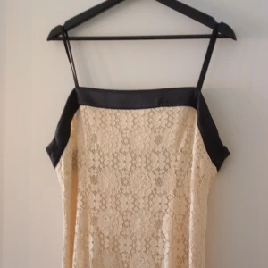 Champagnefärgad spetsklänning med svarta detaljer - Säljer en elegant beige/champagnefärg klänning i spets med svarta detaljer. Klänningen har smala axelband och en stilren design som passar perfekt för festliga tillfällen. Den är i nyskick och har en vacker passform.
