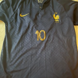 Blå fotbollströja från Nike - Säljer en snygg blå fotbollströja från Nike med guldiga detaljer. Tröjan har korta ärmar och är i storlek XL. Den har nummer 10 och namnet 'MBAPPE' på ryggen. Perfekt för fotbollsfans!