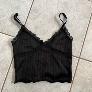 Svart ribbad linne från Divided - Säljer en snygg svart ribbad linne från Divided. Den har spetsdetaljer längs kanterna och justerbara axelband. Perfekt för både vardag och fest! skriv vid frågor💗