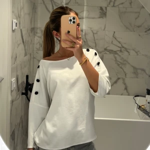 Vit tröja med svarta knappar - Säljer en stilren vit tröja med trekvartsärm och svarta knappar på axlarna. Tröjan har en lös passform och är perfekt för både vardag och fest. Materialet känns mjukt och bekvämt, vilket gör den till ett mångsidigt plagg i garderoben.