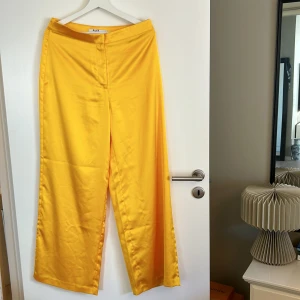 Gula byxor från NA-KD - Säljer ett par supercoola gula byxor från NA-KD. De är högmidjade och har en snygg wide fit som ger en avslappnad men stilren look. Materialet känns som satin, vilket ger en lyxig känsla. Perfekta för både fest och vardag!