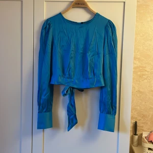 Blå blus från Vero Moda - Säljer en superfin blå blus från Vero Moda. Den har långa ärmar och en snygg knytning i midjan som man knyter bak. Blusen är i ett glansigt material och har en öppen rygg som ger en extra touch. Perfekt för både vardag och fest! kommer med tillhörande byxor om visas på bilden. för båda 120kr och för en del 60kr. super snyggt på.