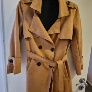Brun trenchcoat med bälte - Säljer en snygg brun trenchcoat med bälte och stora knappar. Den har en klassisk krage och långa ärmar med knappar vid ärmsluten. Perfekt för höst och vår!