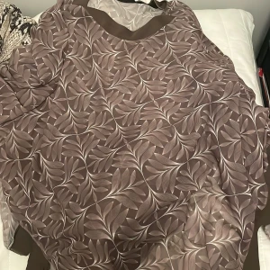 Brun mönstrad klänning från Alfalfa - Säljer en elegant brun klänning från Alfalfa med ett vackert bladmönster. Klänningen har en lös passform och är perfekt för både vardag och fest. Materialet känns lätt och luftigt, vilket gör den bekväm att bära hela dagen.