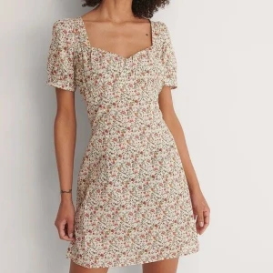 Blommig klänning - Superfin blommig klänning från NA-KD, endast använd vid ett tillfälle så väldigt bra skick. Färgen är krämvit/beige med röda blommor. Den är insydd lite vid brösten vilket syns på sista bilden men det går lätt att sprätta upp.