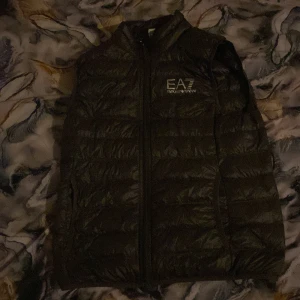 Svart dunväst från EA7 Emporio Armani - Säljer en snygg svart dunväst från EA7 Emporio Armani. Västen har en dragkedja framtill och är perfekt för kyligare dagar. Den är quiltad och ger en stilren look. Passar bra till både vardag och fest.