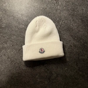 Vit ribbad mössa från Moncler - Säljer en snygg vit ribbad mössa från Moncler i nytt skick. Den är perfekt för vintern och har en broderad logga framtill. Mössan är i one size och gjord av mjukt material som håller dig varm och stilren. Passar både till vardags och mer uppklädda tillfällen. 🧢❄️
