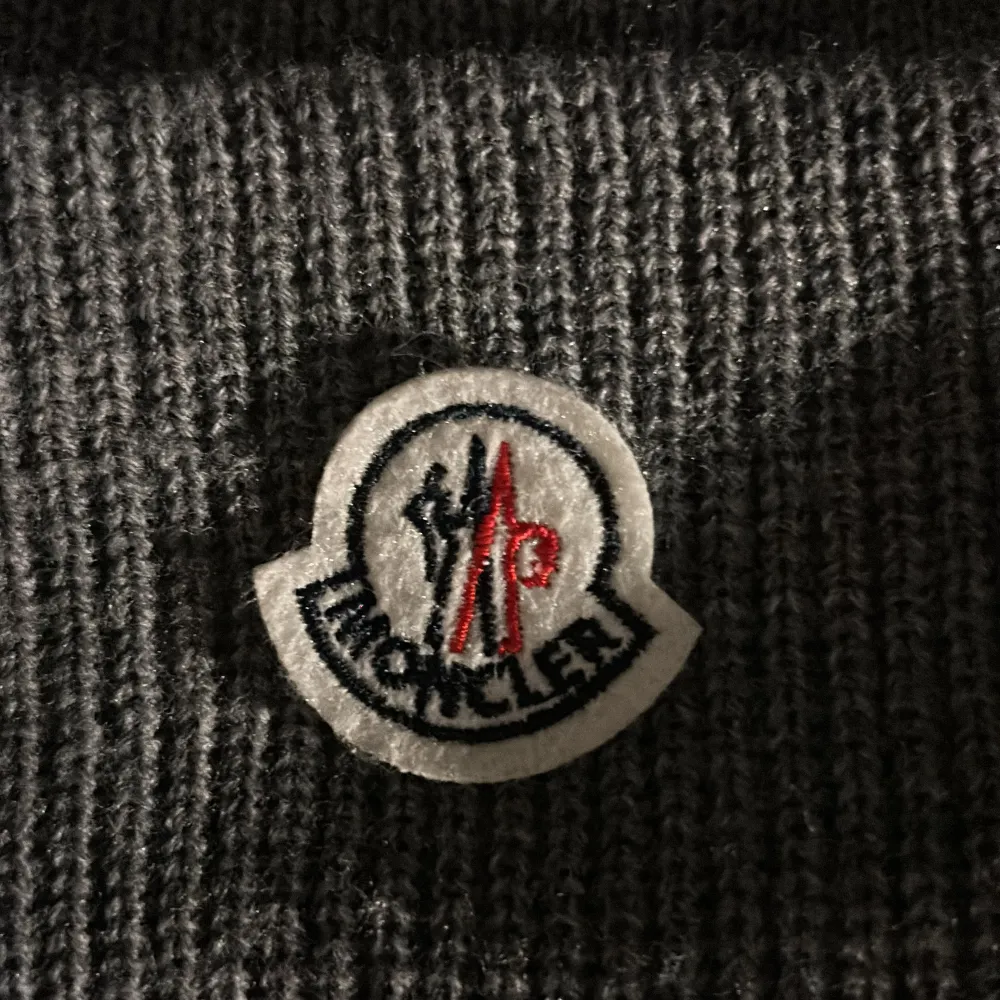 Säljer en snygg grå stickad mössa från Moncler. Den har en broderad logga framtill och är perfekt för kyliga dagar. Mössan är i ett ribbat material som ger en skön passform. Passar både till vardags och mer uppklädda tillfällen.. Accessoarer.