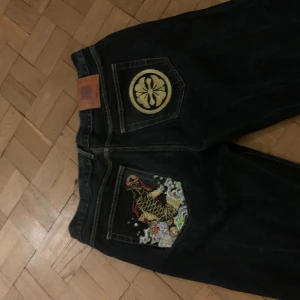 Unika jeans med broderi. Storleken i midjan är 40. Och är ganska baggy  - Säljer ett par coola jeans med unikt broderi på bakfickorna. Ena fickan har ett färgglatt drakmotiv och den andra ett stilrent cirkelmönster. Jeansen är mörkblå och har en klassisk passform med normal midja. Perfekta för dig som vill sticka ut lite extra! 🐉✨