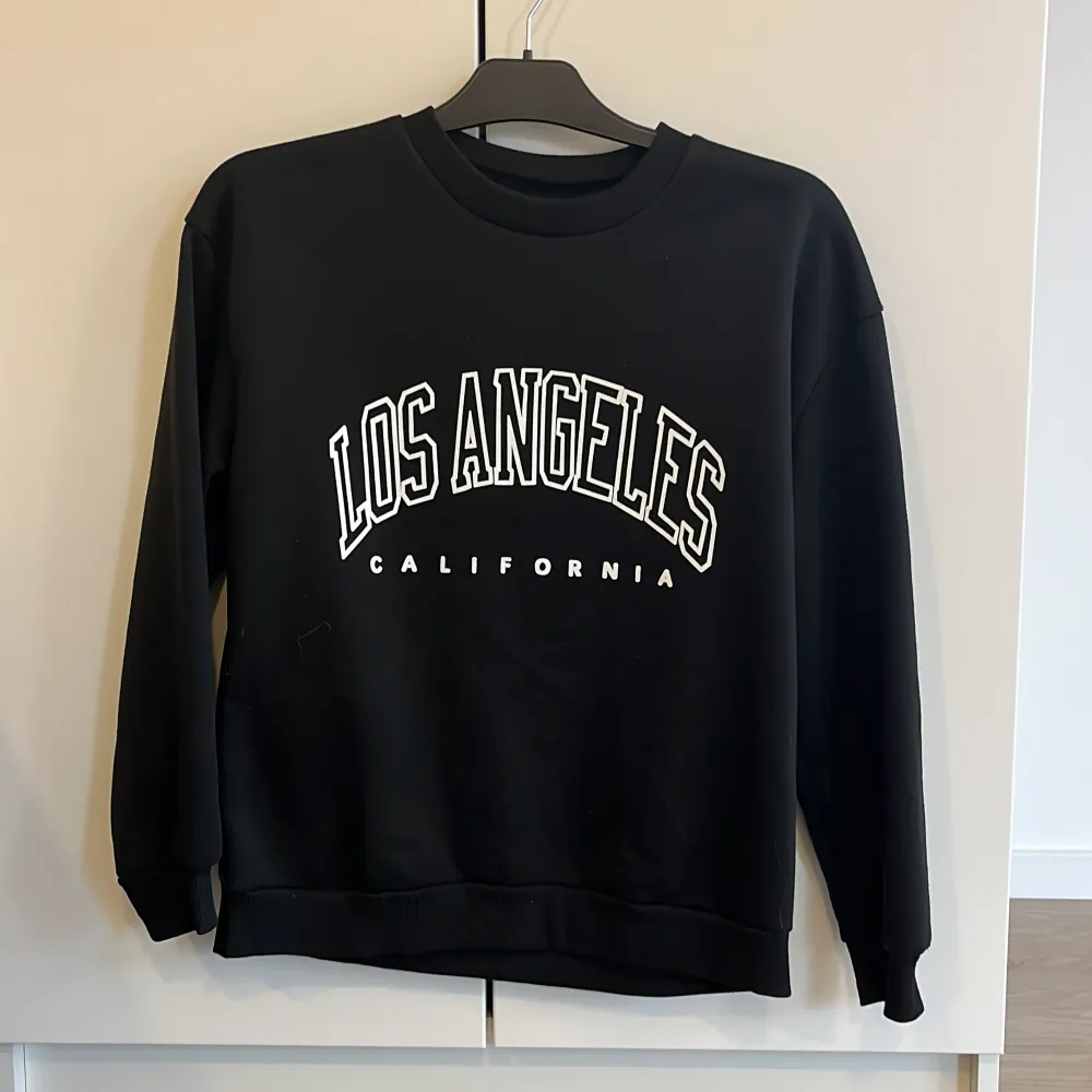 Säljer en svart sweatshirt med texten 'Los Angeles California' i vitt på framsidan. Tröjan har en klassisk rund hals och långa ärmar, perfekt för en avslappnad stil. Den är i bra skick. 🖤. Tröjor & Koftor.