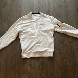 Vit sweatshirt från C.P. Company - Säljer en stilren vit sweatshirt från C.P. Company i storlek M. Tröjan har en cool detalj med en lins på ärmen och är i nytt skick med etikett kvar. Perfekt för både vardag och avslappnade tillfällen. Passar bra till jeans tuvär till det tråkiga märket på armen har gått sönder när jah tvätta den man kan någ köpa ett nytt för en bilig peng den är äkta och köpt på cp companys hemsida 