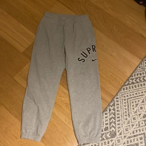 Grå mjukisbyxor från Nike x Supreme - Säljer ett par grå mjukisbyxor från Nike x Supreme. De har en elastisk midja och muddar vid bensluten för en bekväm passform. Stort Supreme-tryck på benen ger en cool look. Perfekta för chill dagar eller streetwear-stil.