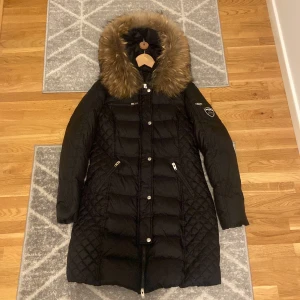 Svart dunjacka från Rockandblue - Säljer en snygg svart dunjacka från Rockandblue i storlek 40. Jackan har en mysig pälsfodrad huva och quiltade detaljer. Perfekt för kalla vinterdagar med sin långa design och dragkedja framtill. Sälj för  mindre användning pga en ny jacka jag skaffat.                                Litet hål vid fickan knappt synlig kunde inte få med på bild, vilket innebär de lilla priset jag lägger ut det för