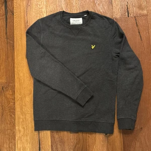 Mörkgrå tröja från Lyle & Scott - Säljer en snygg mörkgrå tröja från Lyle & Scott i ekologisk bomull. Den har en broderad gul logga på bröstet och är perfekt för både vardag och fest. Tröjan har långa ärmar och en klassisk passform. Passar perfekt till höst och vinter! 🍂