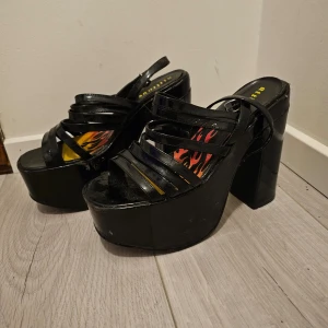 Svarta platåskor med klack - Säljer ett par coola svarta platåskor med hög klack och remmar. De har en innersula med eldflammor i rött och gult. Perfekta för en utekväll eller fest! Skorna är ganska slitna på platåerna. 