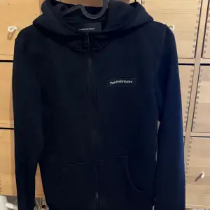 Säljer en stilren svart hoodie från Peak Performance i mycket bra skick, XS ca 12-14 år. Den har en dragkedja framtill och en praktisk huva. Perfekt för både vardag och träning. Stor ficka på magen för extra bekvämlighet.