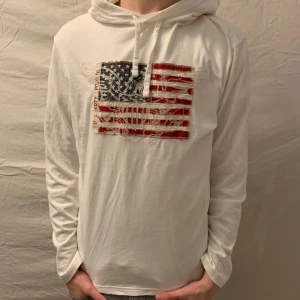 Vit hoodie från Polo Ralph Lauren - Säljer en snygg vit hoodie från Polo Ralph Lauren med ett coolt amerikanskt flaggmönster på framsidan. Den är i mycket bra skick och perfekt för en avslappnad stil. Passar både till vardags och chilliga häng. 🏷️Storlek M