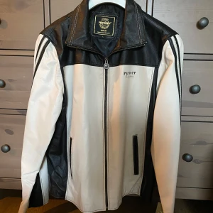 Svart och vit skinnjacka från Petroff - Säljer en snygg svart och vit skinnjacka från Petroff i storlek L. Jackan har en cool dragkedja framtill och stilrena svarta detaljer på ärmarna. Perfekt för höst och vår när du vill ha en stilren look. Den är ny och har lappar kvar på armen. 